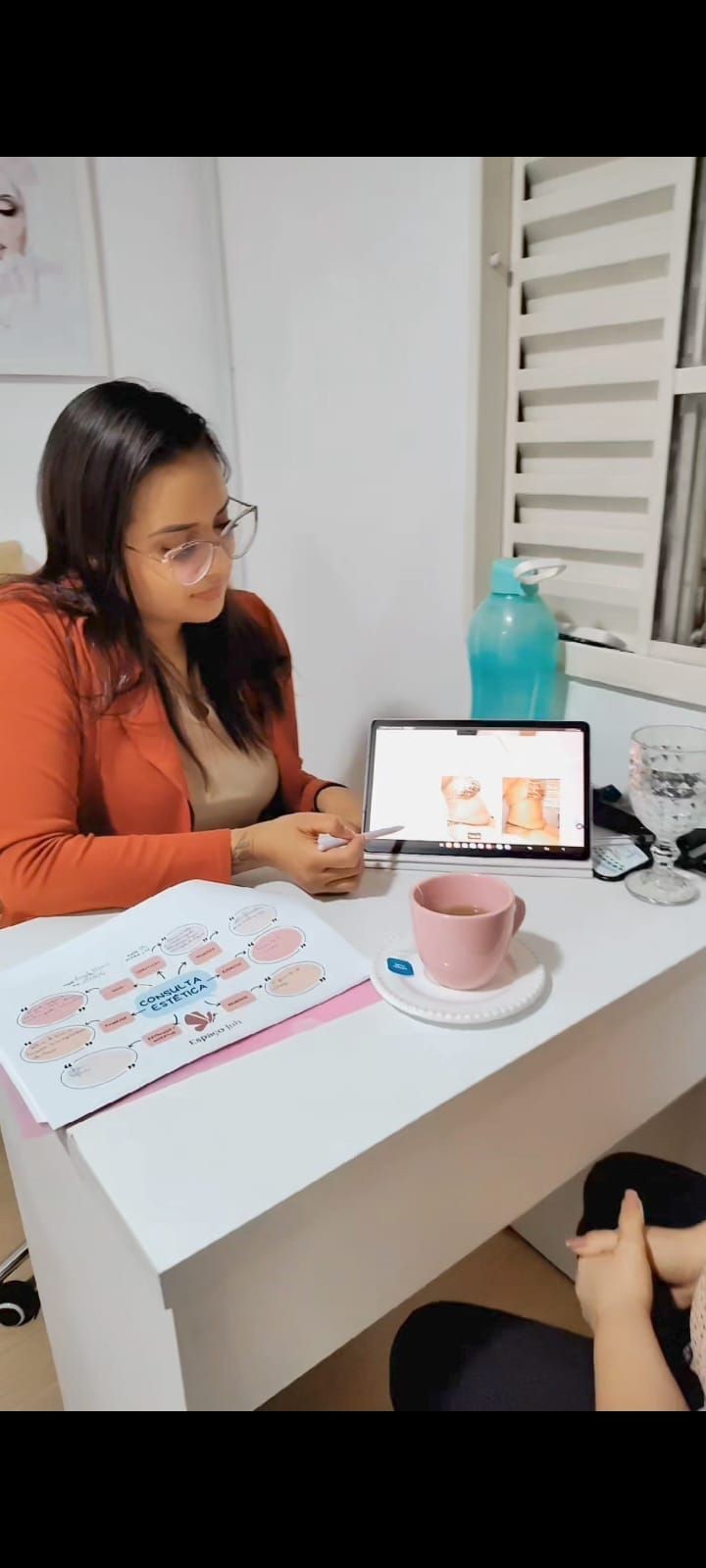 Woman in glasses discussing aesthetics with a client, pointing to images on a laptop at a desk.